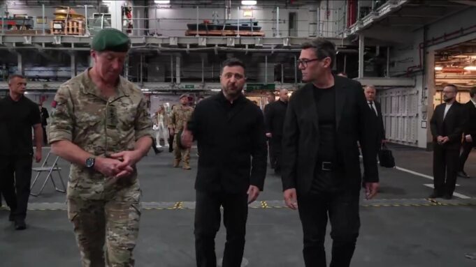 Three men walk through a large industrial hangar: a soldier in camouflage with a green beret, flanked by two men in black outfits, with others in the background.