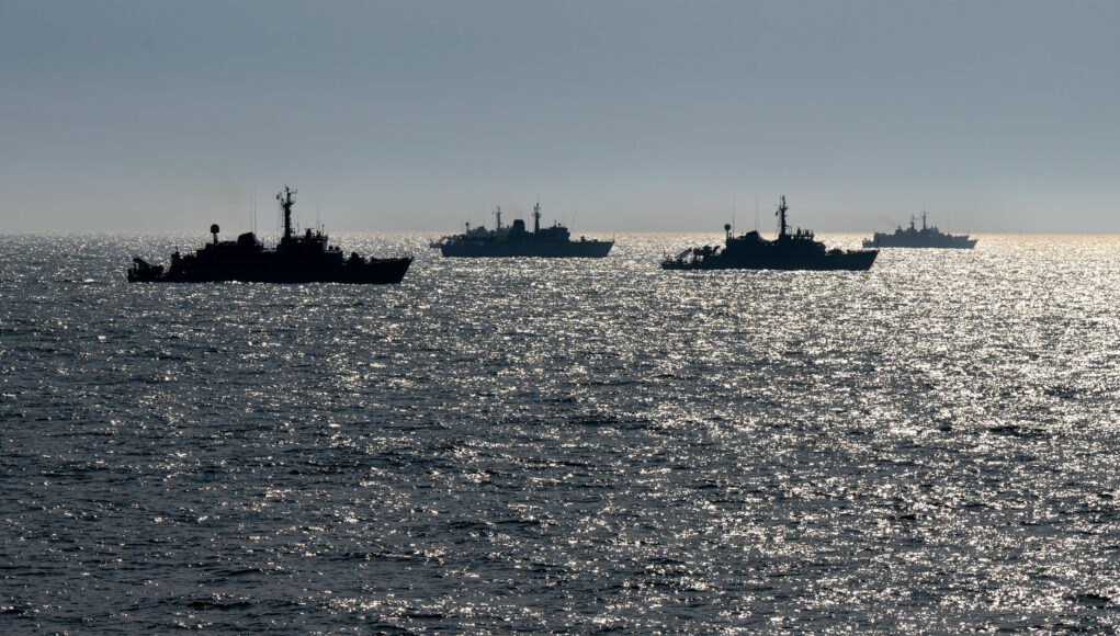 Silhouetted naval ships on a glittering ocean at sunset