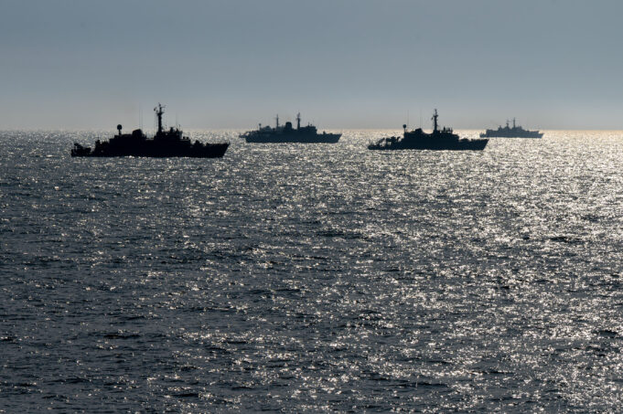 Silhouetted naval ships on a glittering ocean at sunset