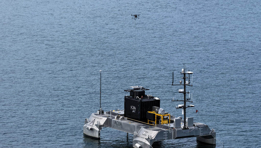 Teledyne TUAS ACUA USV 1 Unmanned research catamaran on calm sea with a black control module and yellow equipment, drone overhead.