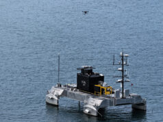 Tethered drone flies from uncrewed vessel in UK demo Unmanned research catamaran on calm sea with a black control module and yellow equipment, drone overhead.