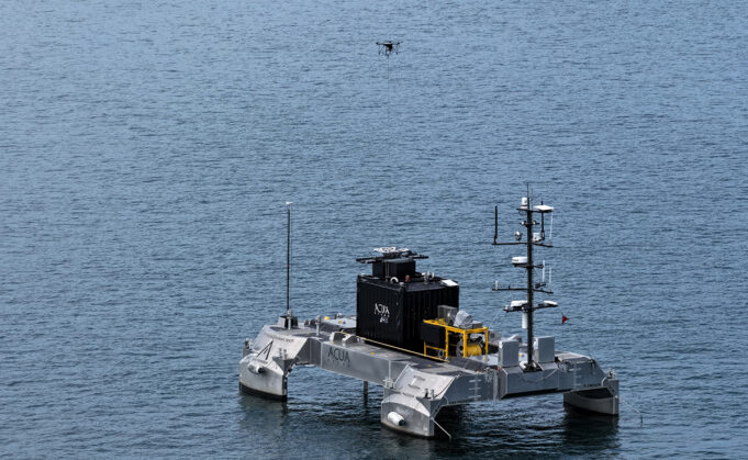Tethered drone flies from uncrewed vessel in UK demo Unmanned research catamaran on calm sea with a black control module and yellow equipment, drone overhead.