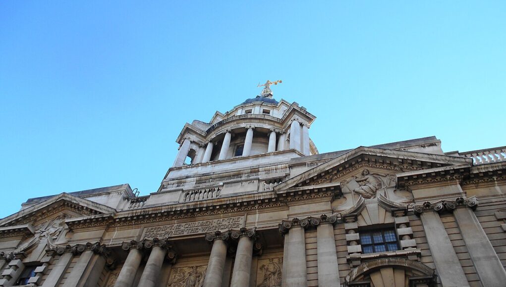 The_Central_Criminal_Court,_Old_Bailey_-_geograph.org.uk_-_5923124 Facade of a grand neoclassical building with tall columns and a domed tower against a blue sky.