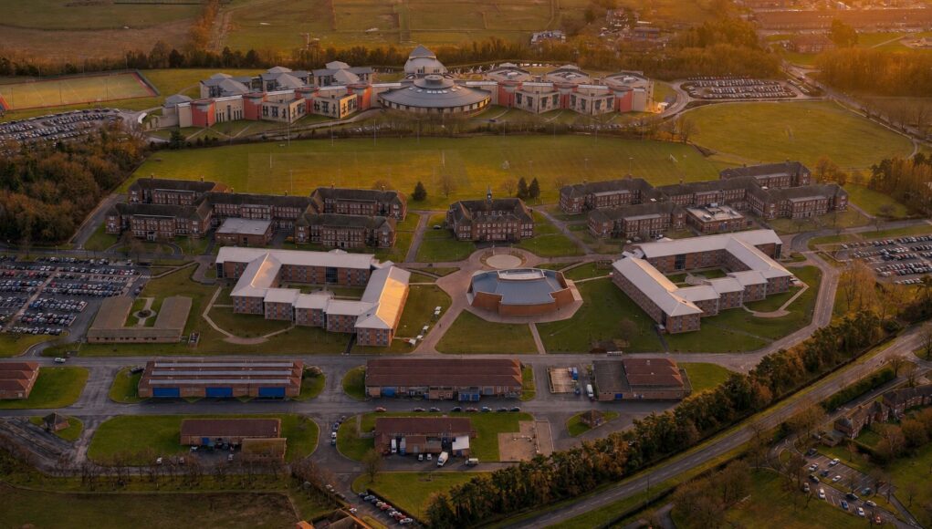 Aerial view of a university campus at sunset: central circular building surrounded by modern brick dorms, athletic fields, and parking lots.