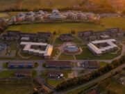 Aerial view of a university campus at sunset: central circular building surrounded by modern brick dorms, athletic fields, and parking lots.