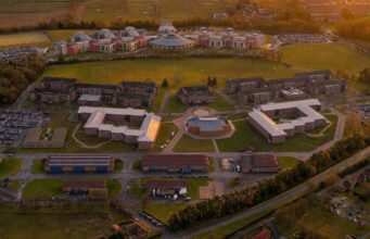 Man jailed over reconnaissance at UK military academy Aerial view of a university campus at sunset: central circular building surrounded by modern brick dorms, athletic fields, and parking lots.