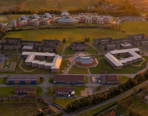 Aerial view of a university campus at sunset: central circular building surrounded by modern brick dorms, athletic fields, and parking lots.