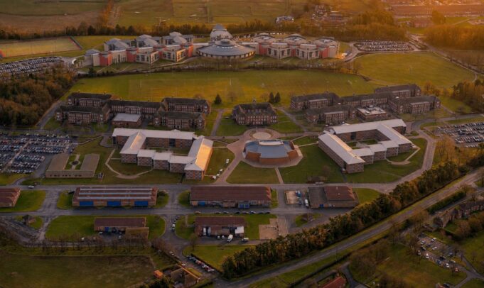 Aerial view of a university campus at sunset: central circular building surrounded by modern brick dorms, athletic fields, and parking lots.