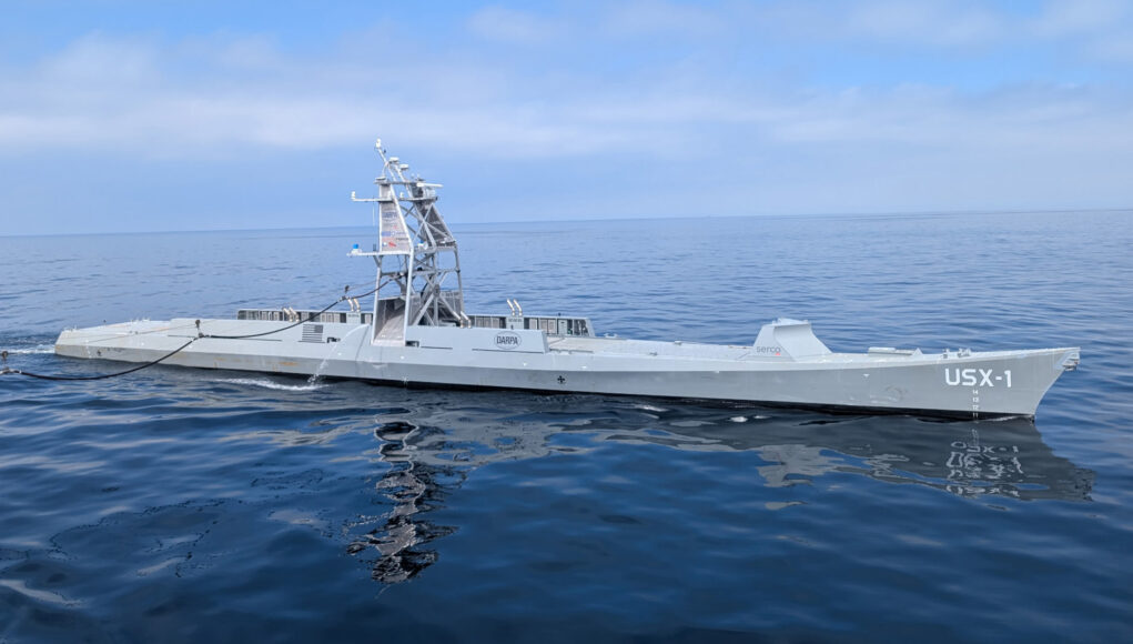 USX-1 Defiant Large gray naval vessel with a tall lattice-like superstructure sailing on calm blue ocean.”