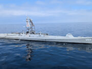 Large gray naval vessel with a tall lattice-like superstructure sailing on calm blue ocean.&rdquo;
