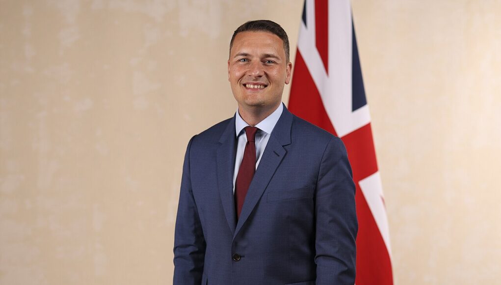 Wes_Streeting_Official_Cabinet_Portrait,_July_2024 Smiling man in a blue suit and red tie standing in front of a British flag.