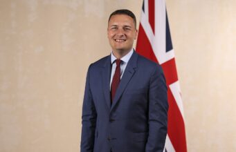 Wes Streeting appointed Defence Secretary Smiling man in a blue suit and red tie standing in front of a British flag.