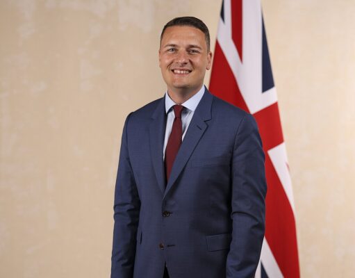 Wes Streeting appointed Defence Secretary Smiling man in a blue suit and red tie standing in front of a British flag.