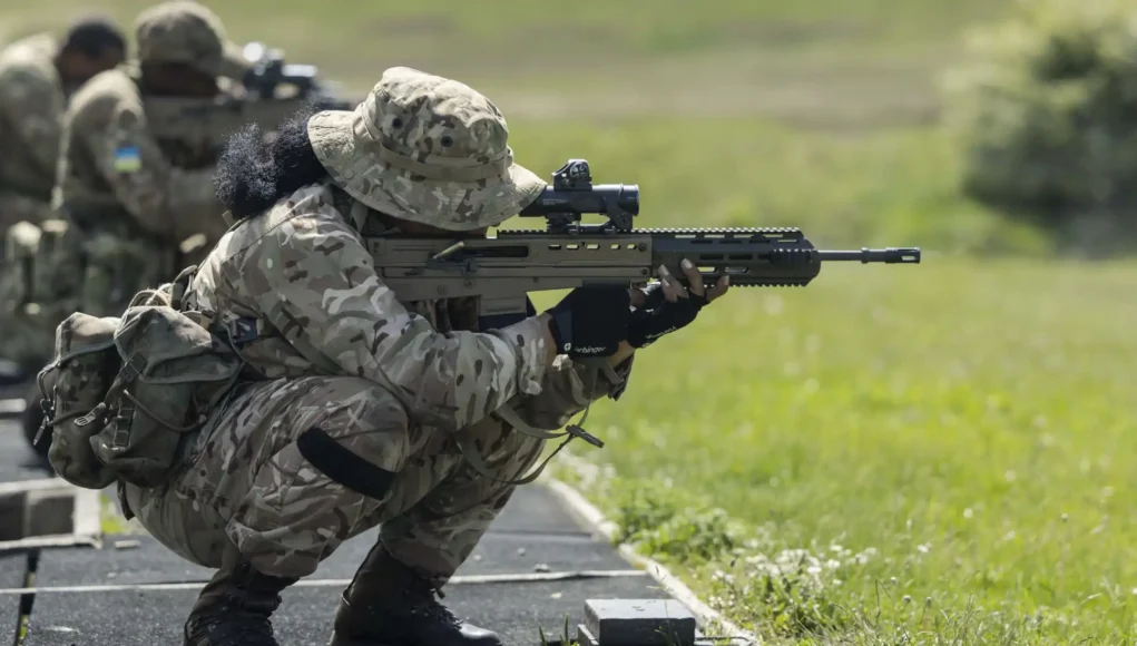 Soldier in camouflage crouches and aims a long rifle during a training exercise on a grassy range at the edge of a paved path.