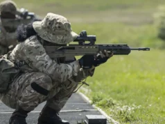 Royal Bermuda Regiment deploys to UK for two-week exercise Soldier in camouflage crouches and aims a long rifle during a training exercise on a grassy range at the edge of a paved path.