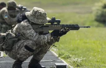 Royal Bermuda Regiment deploys to UK for two-week exercise Soldier in camouflage crouches and aims a long rifle during a training exercise on a grassy range at the edge of a paved path.