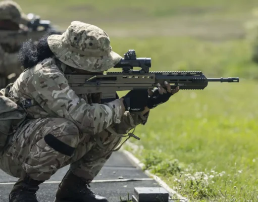 Royal Bermuda Regiment deploys to UK for two-week exercise Soldier in camouflage crouches and aims a long rifle during a training exercise on a grassy range at the edge of a paved path.