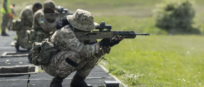 Soldier in camouflage crouches and aims a long rifle during a training exercise on a grassy range at the edge of a paved path.
