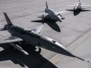 Three military fighter jets parked on a concrete airstrip with their shadows visible on the tarmac, seen from above.