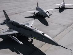Three military fighter jets parked on a concrete airstrip with their shadows visible on the tarmac, seen from above.