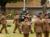 Group of soldiers in brown dress uniforms marching with large artillery guns in the background outdoors.