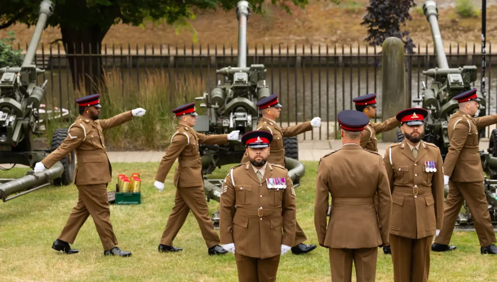 army031-official-20260717-0014 Group of soldiers in brown dress uniforms marching with large artillery guns in the background outdoors.