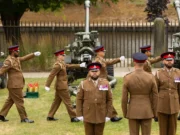 Group of soldiers in brown dress uniforms marching with large artillery guns in the background outdoors.