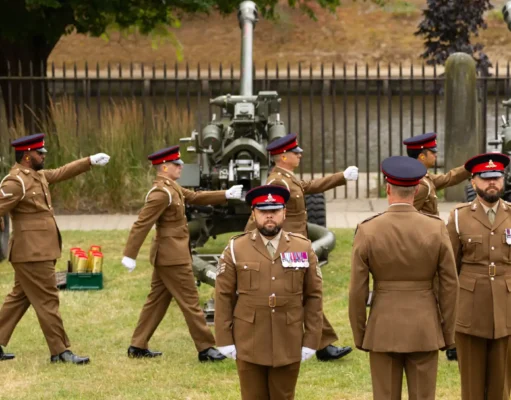 Group of soldiers in brown dress uniforms marching with large artillery guns in the background outdoors.