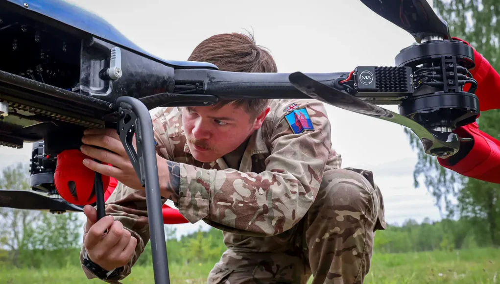 army900-official-260522-001-0013 Military technician in camouflage crouches under a large unmanned aerial vehicle, inspecting its underside in a grassy field.