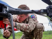 British Army tests loitering munitions in Estonia Military technician in camouflage crouches under a large unmanned aerial vehicle, inspecting its underside in a grassy field.