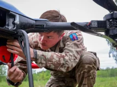 Military technician in camouflage crouches under a large unmanned aerial vehicle, inspecting its underside in a grassy field.