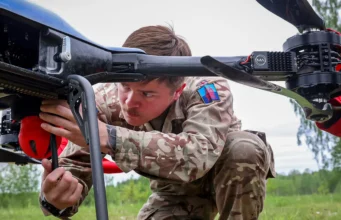 British Army tests loitering munitions in Estonia Military technician in camouflage crouches under a large unmanned aerial vehicle, inspecting its underside in a grassy field.