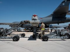 Ground crew service a large military aircraft on a tarmac with loading equipment and vehicles nearby under a clear blue sky.