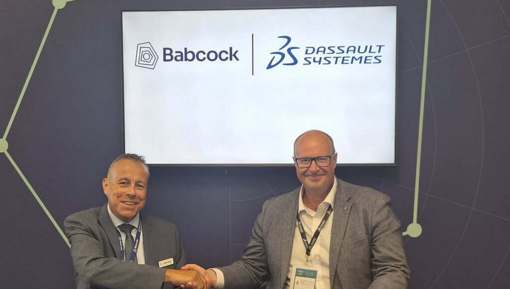 Two men in suits shake hands across a table during a signing ceremony, with Babcock and Dassault Syst&egrave;mes logos displayed on a screen behind them.