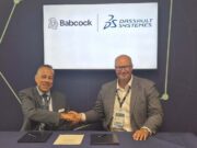 Two men in suits shake hands across a table during a signing ceremony, with Babcock and Dassault Syst&egrave;mes logos displayed on a screen behind them.
