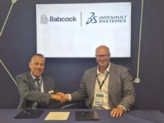 Two men in suits shake hands across a table during a signing ceremony, with Babcock and Dassault Syst&egrave;mes logos displayed on a screen behind them.