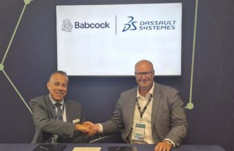 Two men in suits shake hands across a table during a signing ceremony, with Babcock and Dassault Syst&egrave;mes logos displayed on a screen behind them.
