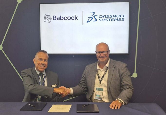 Two men in suits shake hands across a table during a signing ceremony, with Babcock and Dassault Syst&egrave;mes logos displayed on a screen behind them.