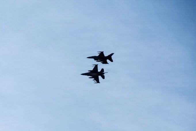 Three silhouetted fighter jets fly in formation across a clear blue sky, with faint reflections below on the water surface.