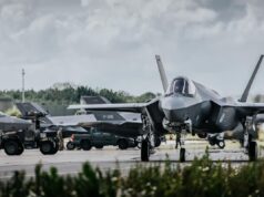 Front view of a stealth fighter on a runway at a military airbase, with ground crew and support vehicles in the background.