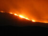 Fire rips along a hillside at night, bright orange flames lighting the sky and smoke.