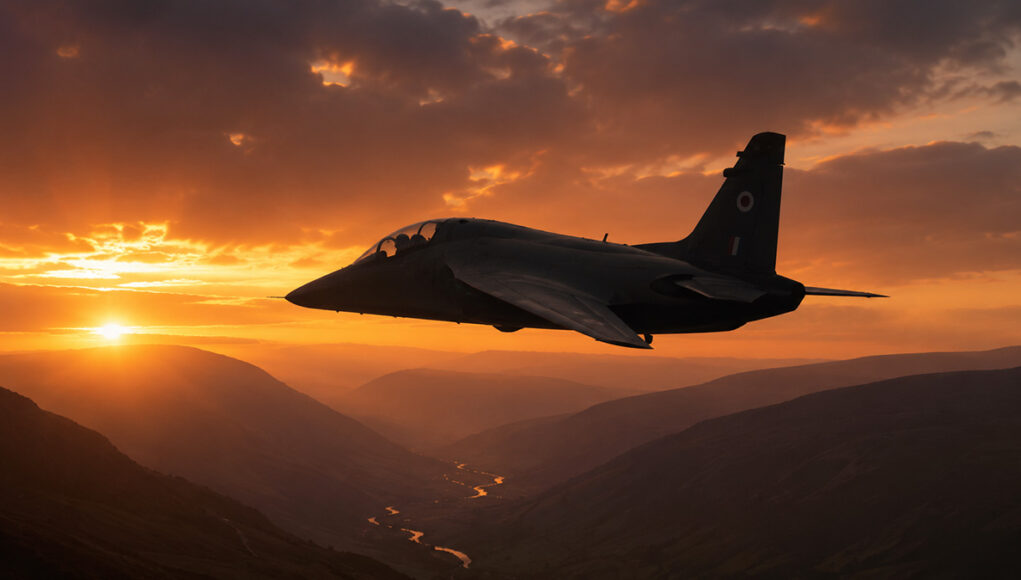 Two-seat military trainer jet silhouetted against a glowing orange sunset over a winding river and mountainous valleys.