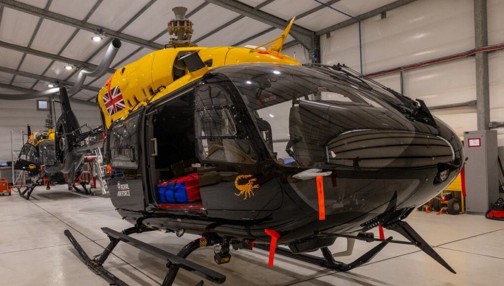 Military helicopters in a spacious hangar; a black helicopter in the foreground with a bright yellow top and a Union Jack emblem on its side