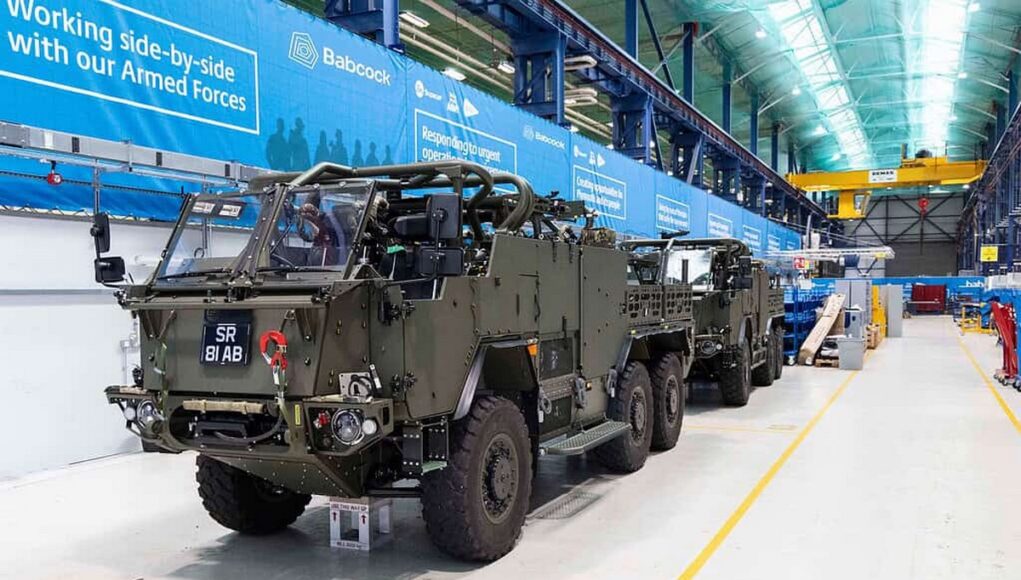 Armored military vehicles lined up inside a large warehouse or maintenance hangar with blue banners along the wall and yellow safety lines on the floor.