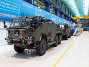 Final Jackal 3 rolls off the line for the British Army Armored military vehicles lined up inside a large warehouse or maintenance hangar with blue banners along the wall and yellow safety lines on the floor.