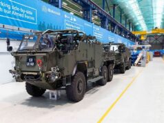 Final Jackal 3 rolls off the line for the British Army Armored military vehicles lined up inside a large warehouse or maintenance hangar with blue banners along the wall and yellow safety lines on the floor.