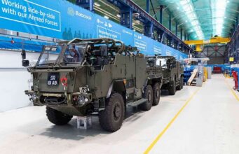 Final Jackal 3 rolls off the line for the British Army Armored military vehicles lined up inside a large warehouse or maintenance hangar with blue banners along the wall and yellow safety lines on the floor.
