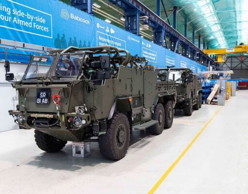 Final Jackal 3 rolls off the line for the British Army Armored military vehicles lined up inside a large warehouse or maintenance hangar with blue banners along the wall and yellow safety lines on the floor.