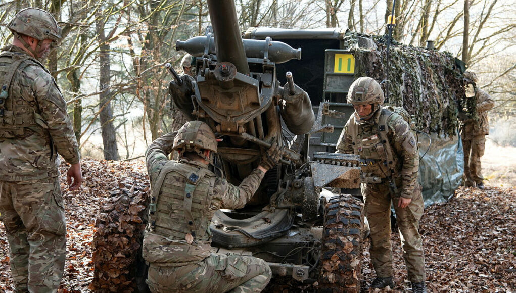 Soldiers in camouflage work on a large artillery piece in a leaf-covered forest.