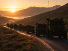 Convoy of armored military vehicles driving along a mountain road at sunset, silhouetted against a golden valley.